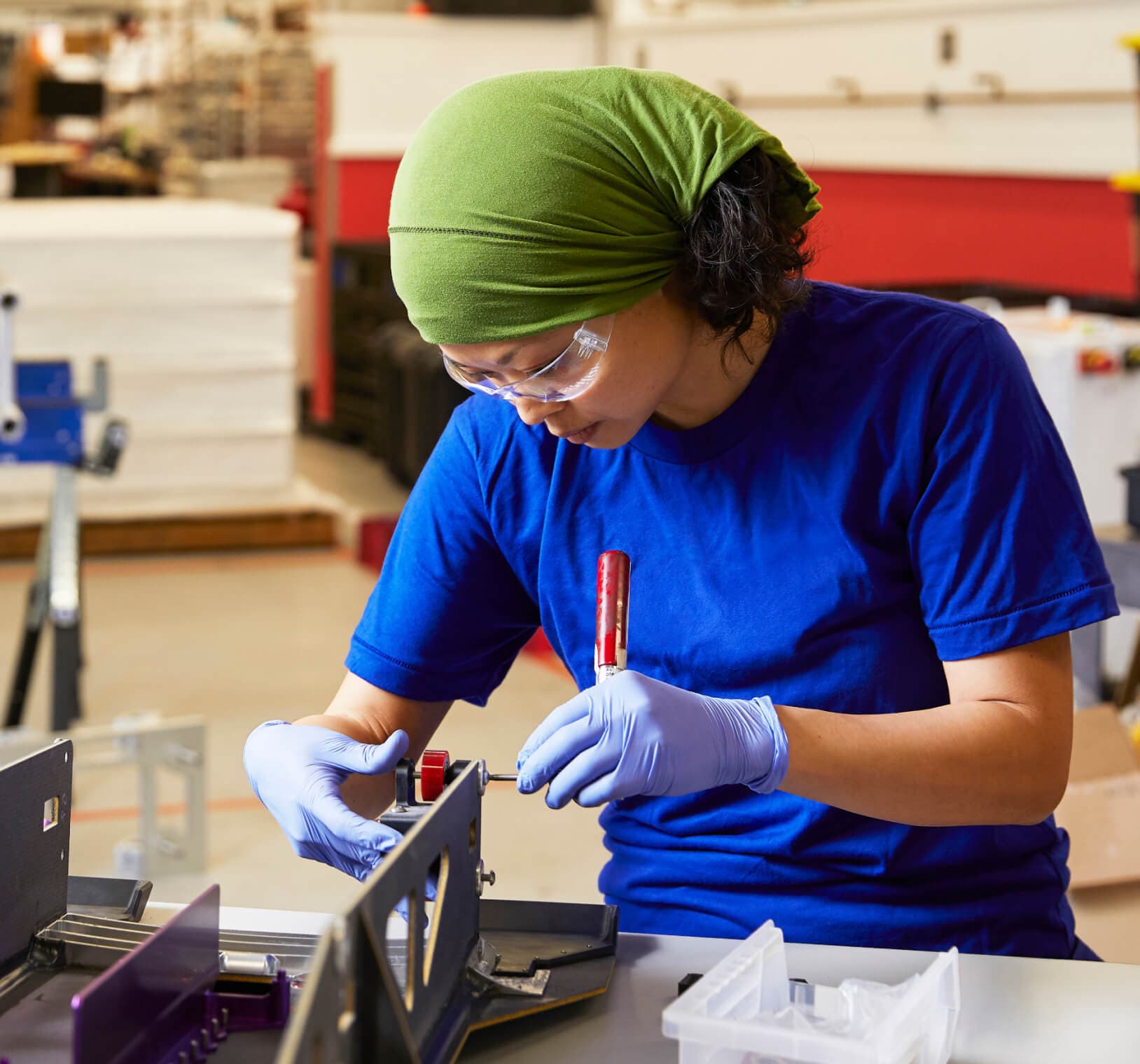 Woman wearing safety glasses and latex gloves carefully assembling a metal component with a screwdriver on a workbench in a manufacturing facility.