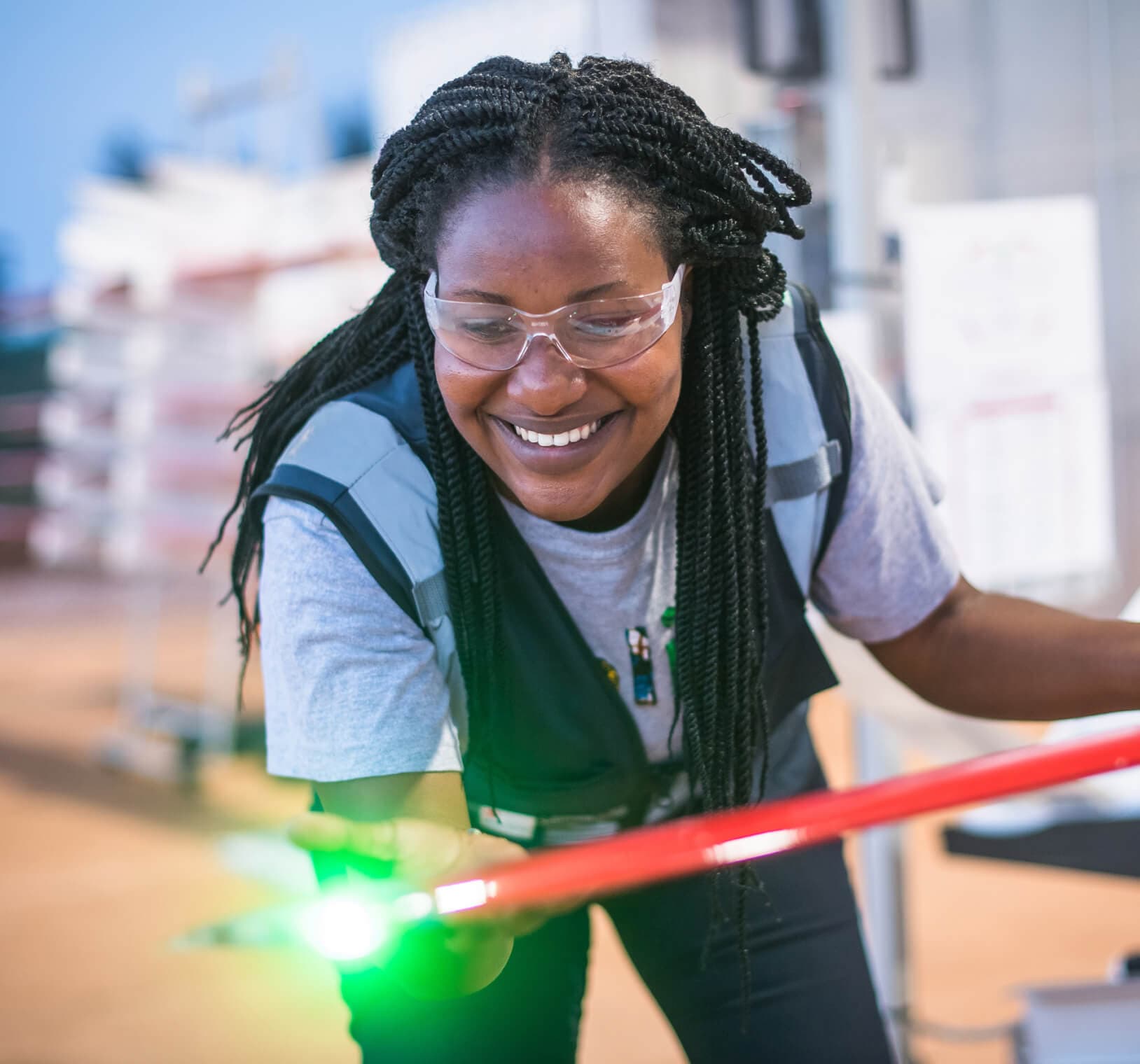 Woman wearing safety glasses and work vest smiling while inspecting a drone component in a manufacturing facility.