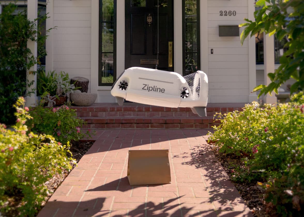 Zipline drone hovering above a delivered package at a house entrance.