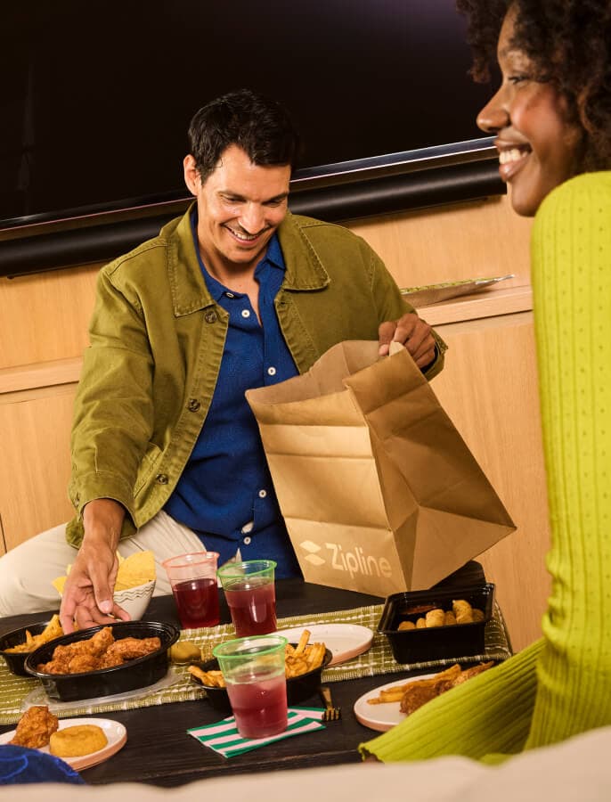 Two people unpacking a Zipline delivery bag at a table with food.