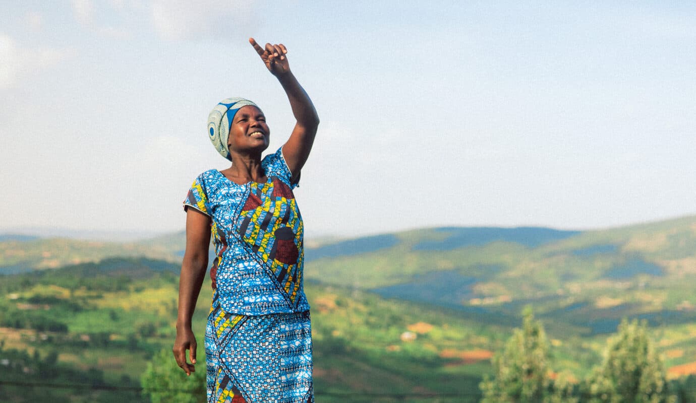 A woman stands on a hillside in Rwanda, looking up and pointing at the sky.